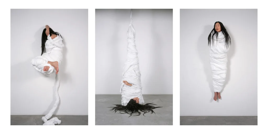 Three images showing a woman with black hair bound in a white sheet in three different positions.