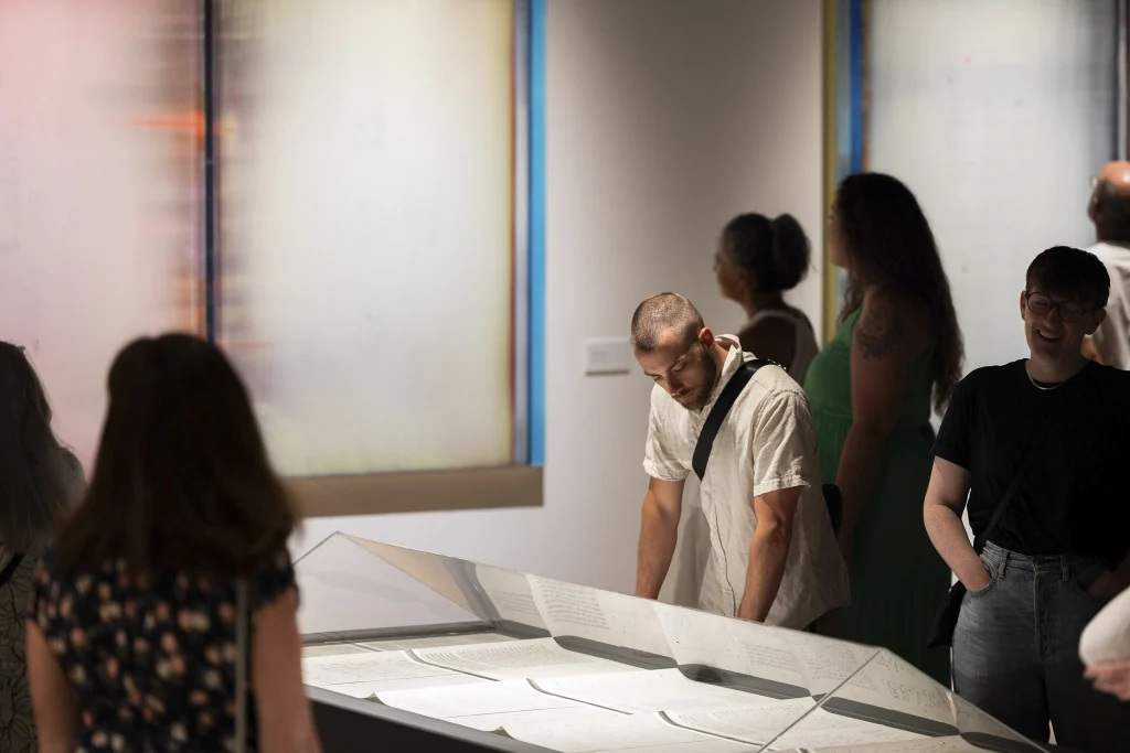 People view Alice Teichert's exhibition. In the center of the image is a plinth with a triangular plexiglass top. A person looks down at the artwork on display