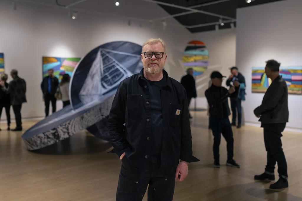 Artist Stan Olthuis stands in the gallery during his opening reception. Slightly out of focus in the background is a large scale kinetic sculpture and people mingling and viewing Stan's artwork