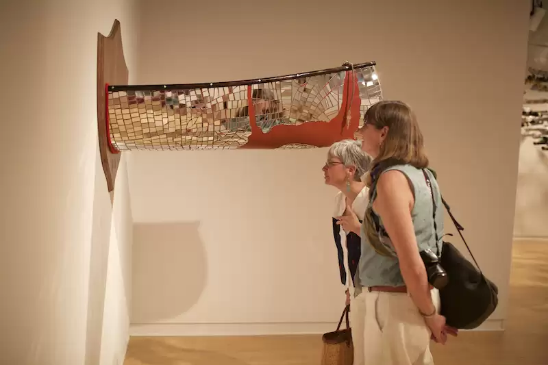 two people view an installation based artwork of a mirror mosaic canoe cut in half and mounts on a plaque on a gallery wall
