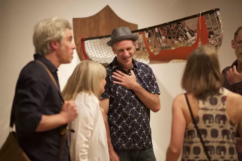 Brad Copping stands in front of one of his sculptural installation pieces with a hand on his chest, smiling at a gathering of people around him