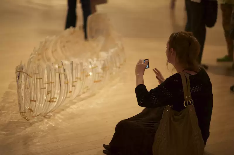 a person sitting on the ground and holding up their phone on the right to take a photo of a glass canoe on the left