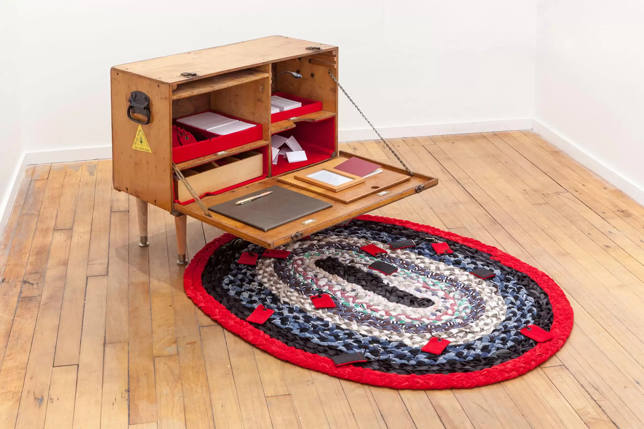 Sculpture by Tim Whiten. Archival wooden box with stationary supplies. Weaved fabric rug with red and black fabric tags.