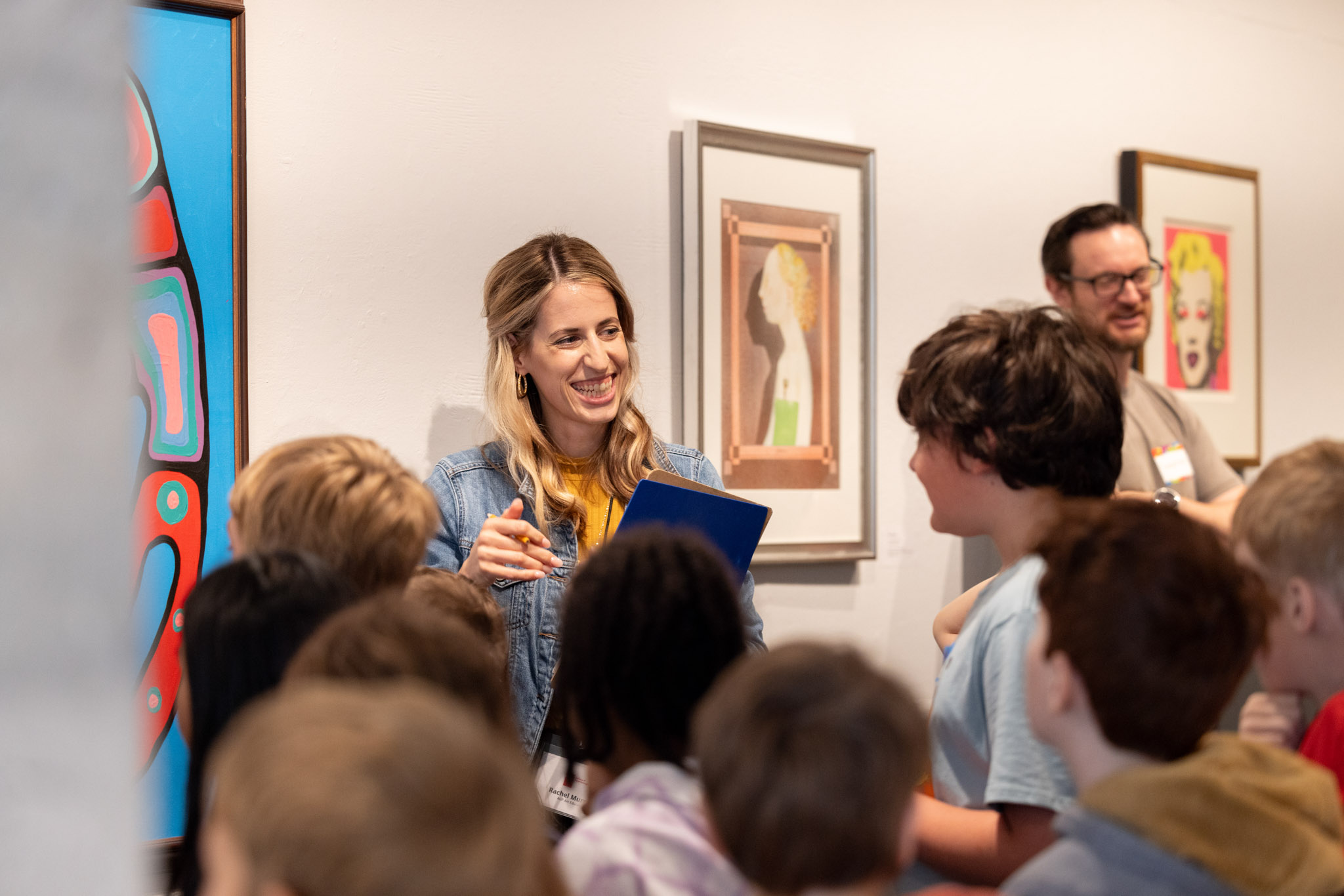 a teacher stands in front of paintings in the gallery and smiles at a group of students who are all facing her with their backs to the camera