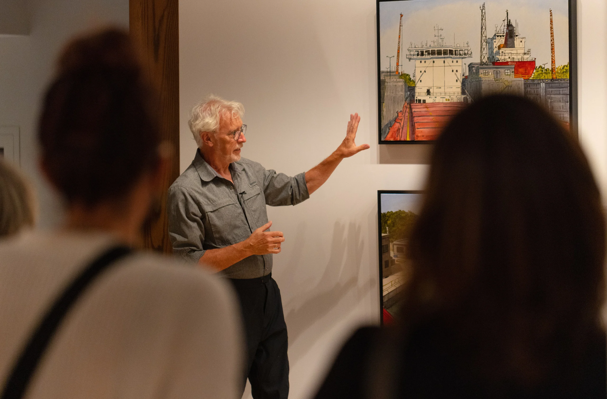 artist Peer Christensen holds a hand up while looking at one of his paintings in the gallery. In the foreground two people are pictured from behind, facing Peer