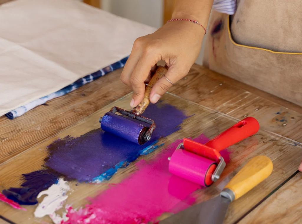 A close up view of a person holding a brayer covered in indigo coloured ink. On the table to the right is another brayer covered in magenta ink