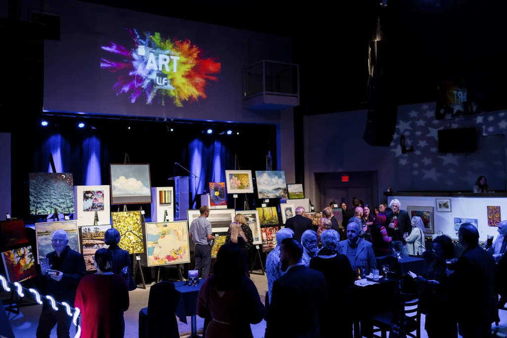 A room full of people and artwork on display at the It's All about ART auction. The event logo appears on a screen above the crowd and a display of artwork extends from the left toward the center of the image