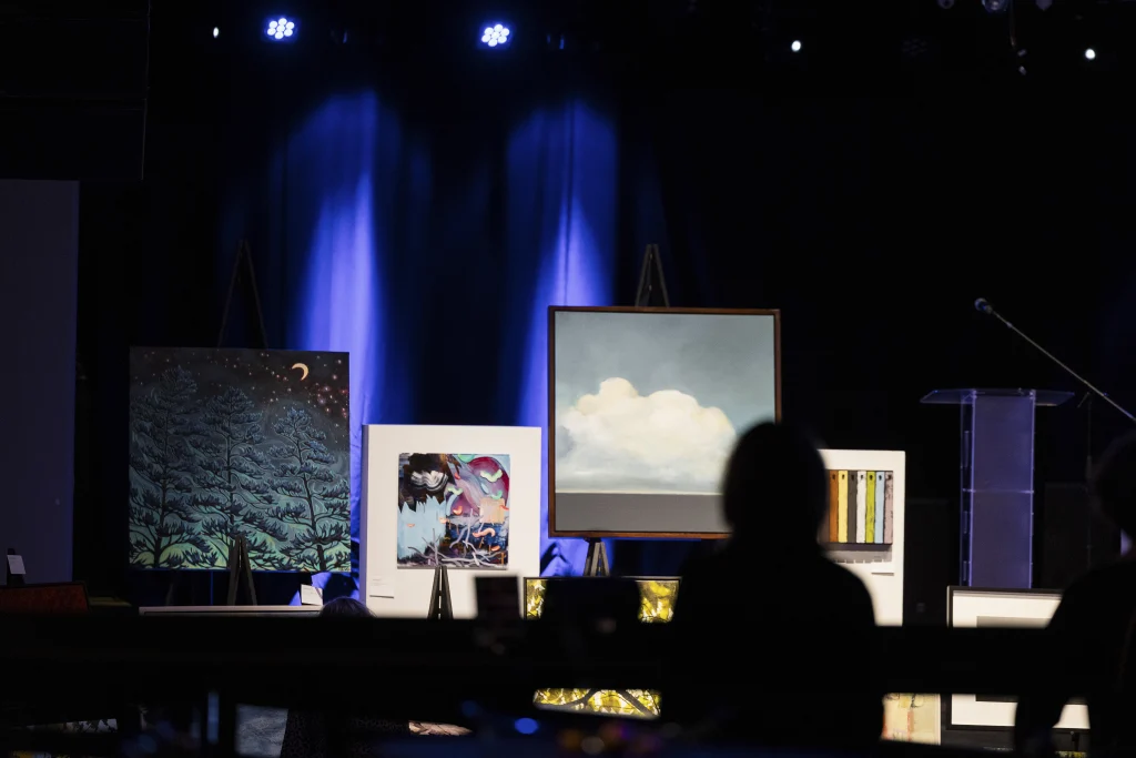 A display of artwork is lit in a dark room. In front is a dark silhouette of a person viewing the artwork