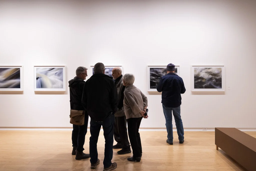 A group of visitors chat amongst themselves in Arnold Zageris' solo exhibition "Refractions." In the background five photographs hang on the wall of the gallery