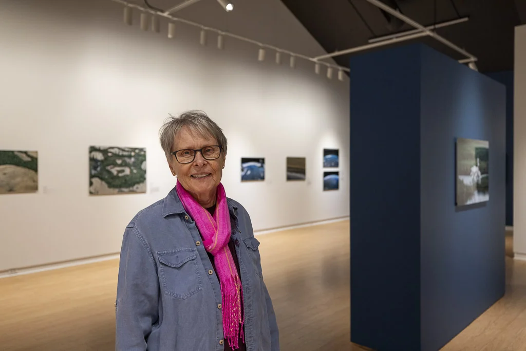 Roberta Bondar stands in her solo exhibition wearing a blue shirt and bright pink scarf