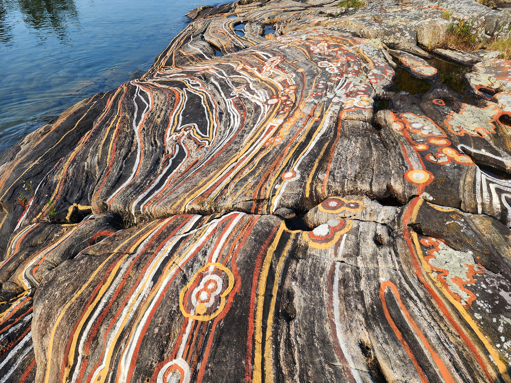 Rock face mural by the artist Cole Swanson with swirling orange, white, and yellow lines by a calm blue water body.