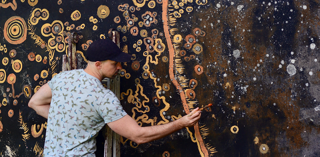 Cole Swanson wearing a patterned shirt and black cap paints an abstract design of orange and beige circles and lines on a rockface.