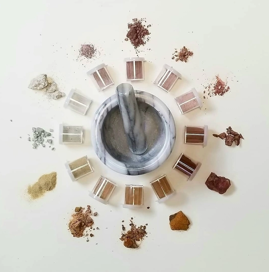 Marble mortar and pestle centered amidst samples of clay, pigments, and small containers arranged in a circle, prepared by the artist Cole Swanson