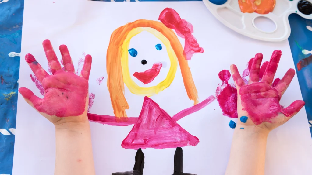 Child's painting of a smiling girl with orange hair in a pink dress. Two small hands with pink paint are imprinted on the paper. Playful and creative scene.