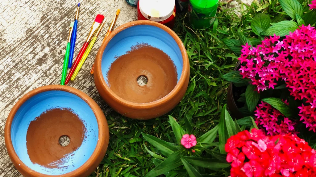 Clay bowls with blue interiors sit on grass beside vibrant pink flowers. Paintbrushes and paint jars are nearby on a textured surface, suggesting a creative, lively atmosphere.