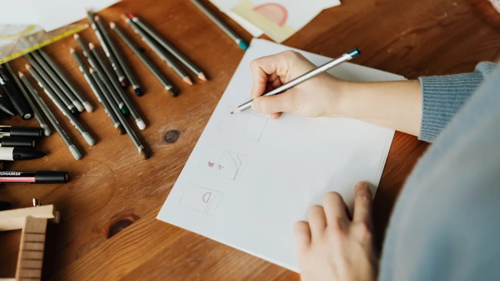A person sketches designs on a blank sheet of paper with a pencil, surrounded by scattered art supplies on a wooden table, conveying creativity and focus.