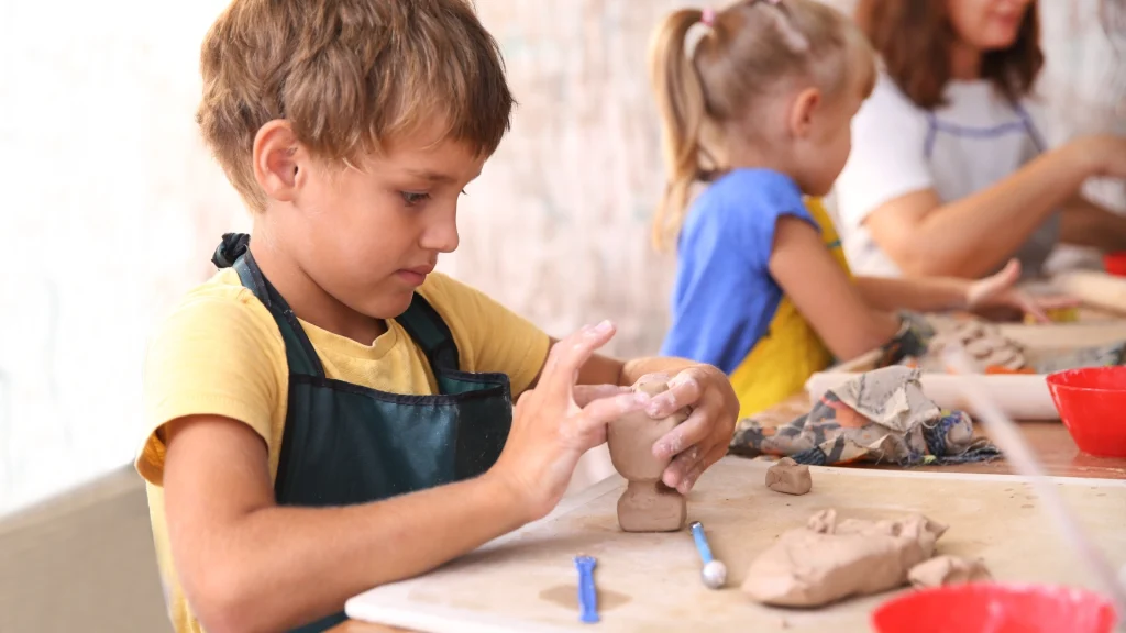 A young boy in a yellow shirt and apron is focused on molding clay at a table. Other children and an adult are in the background, in a creative, educational setting.