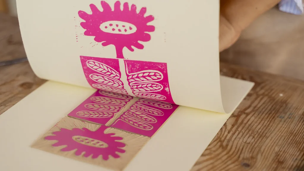 A person lifts paper revealing a pink, handcrafted ink print of a stylized flower with a geometric pattern on a wooden table. The scene is artistic and creative.