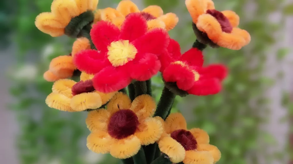 A bouquet of handmade flowers made from red, yellow, and orange pipe cleaners with green stems, set against a blurred green background.