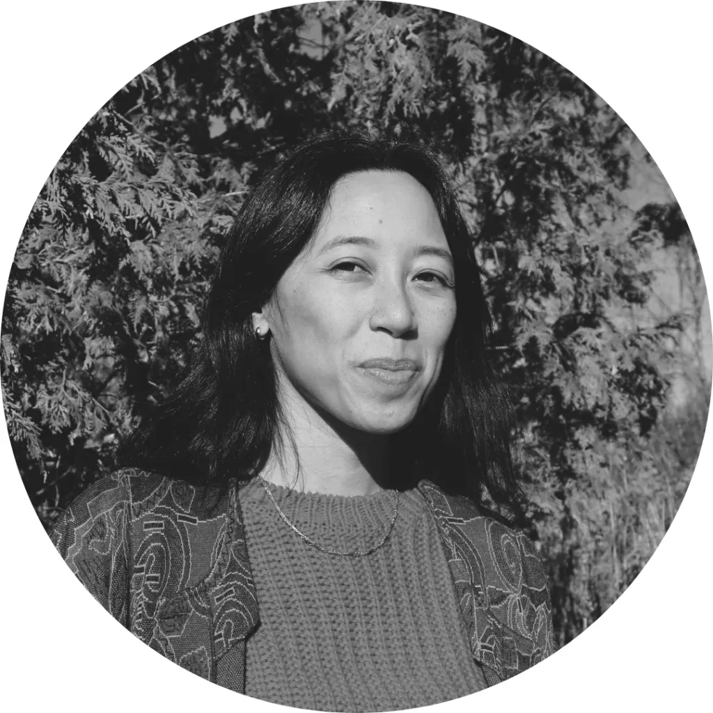 Casandra Lee smiling with long dark hair standing outdoors in front of leafy trees. She wears a patterned jacket and knit sweater. 