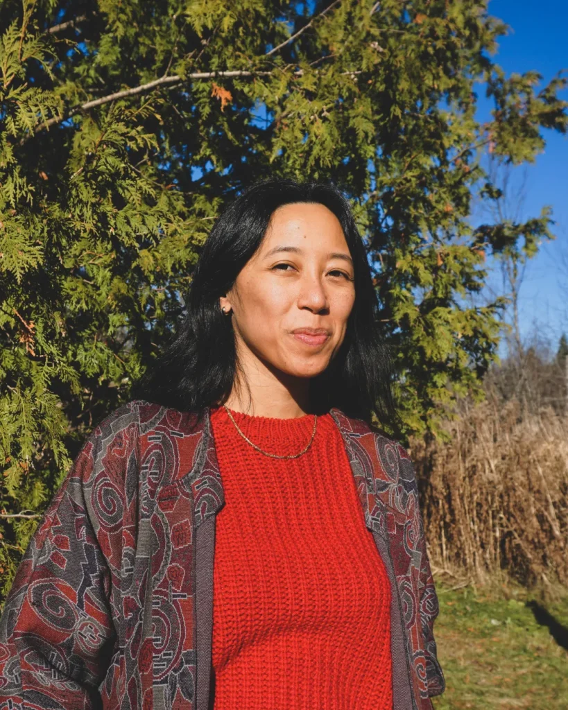 Casandra Lee smiling with long dark hair standing outdoors in front of leafy trees. She wears a patterned jacket and red knit sweater.