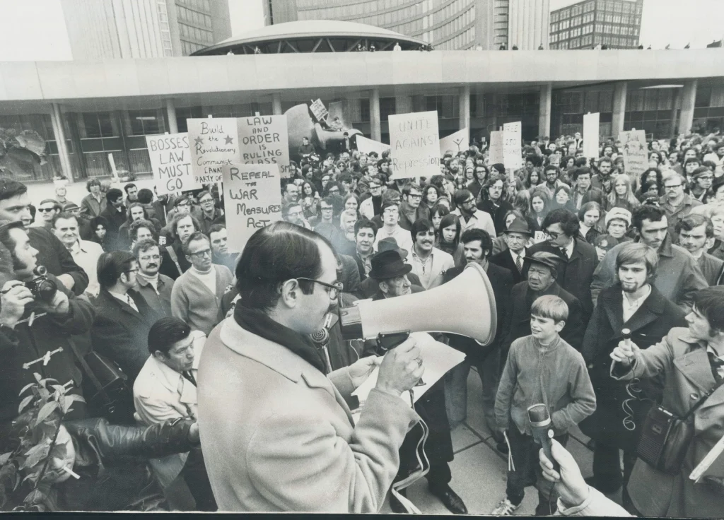 Black and white photograph of a large protest gathering with a man speaking through a megaphone to a crowd holding signs advocating for law and order, repealing war measures, and government accountability. Signs include messages like "BOSSES LAW MUST GO" and "LAW AND ORDER RULES."
