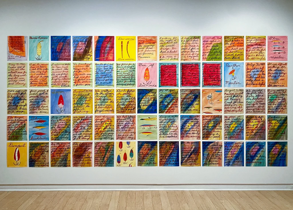 Photograph of a large wall installation by Dennis Tourbin composed of 56 colorful square panels arranged in seven rows and eight columns, each featuring handwritten text and abstract shapes in varied colors like red, blue, yellow, and orange. The panels display a mix of cursive writing and simple illustrations