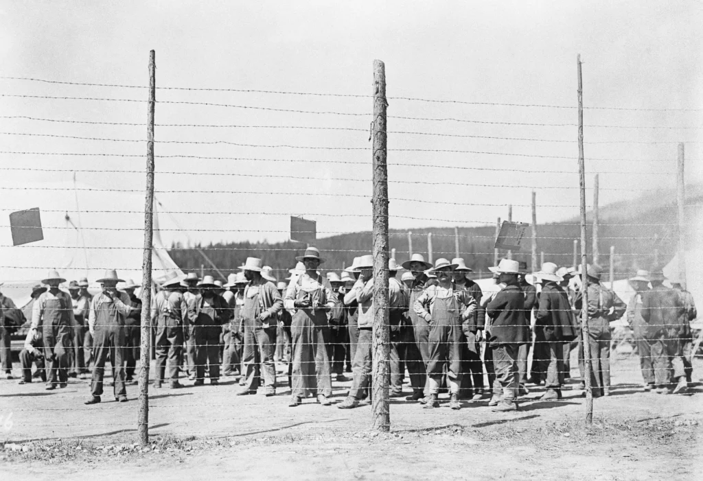 Black and white photograph showing a group of men standing behind a barbed wire fence, wearing hats and work clothes. Wooden posts support the fence, and tents or temporary structures are visible in the background.