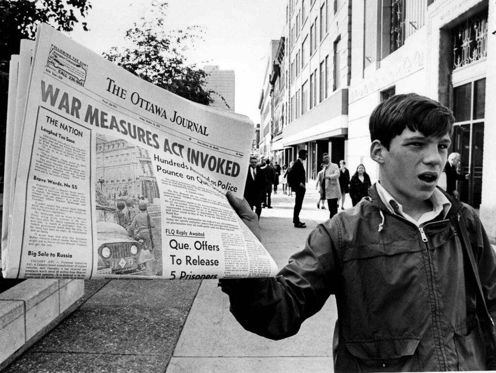 Black and white photograph showing a young person on a city street holding up a newspaper with the headline "War Measures Act Invoked."