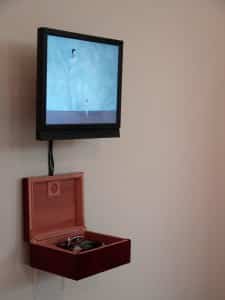 Digital and mixed media installation by Michelle Gay. Screen featuring two people dressed in white and a small open wooden box filled with technological hardware installed on a white wall