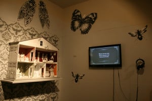 Installation by Jennifer Linton featuring a dollhouse, TV screen with headphones, and and large insect drawings installed on a wall
