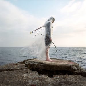 Photograph by Meryl McMaster featuring a person standing on a rocky shore holding a bow and arrows and covered in clear plastic