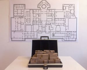Sculpture by Jeannie Thib. Black and white blueprint of a large building installed on a white wall. Open suitcase filled with block like forms installed on a white plinth.