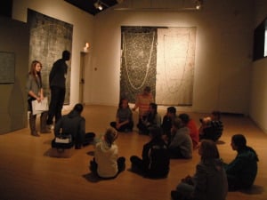 Group of children sitting cross legged discussing various textile works by Dorthy Caldwell installed in AGP's main gallery.
