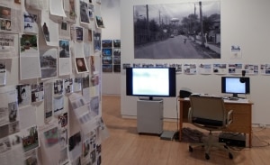 Gallery view of the exhibition Erratic Space. Two screens, a desk, and various articles and photographs installed in AGP's main gallery space.