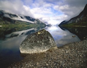 Photograph by Arnold Zageris featuring a large rock on a shoreline. Still water and white clouds in a blue sky.