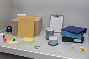Sculpture by Roula Partheniou installed on a white shelf. Series of office supplies without labels.