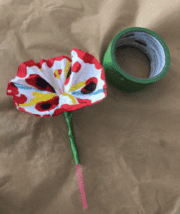 Paper flower dyed with red, blue, and yellow watercolour, pink straw as a stem attached with green painter's tape. Flower is laying on a table beside a roll of green painter's tape.