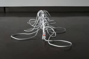 Michelle Bellemare's plymer clay and sculpture of an elongated power cord with several cords plugged in to its outlets. Red light is on.