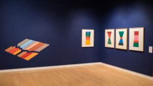 Gallery view of the exhibition duet featuring four abstract block prints by Jack Bush and a digital drawing installed as vinyl by Francisco-Fernando Granados. Prints and digital drawing are mounted on a navy blue wall in the AGP’s main gallery.