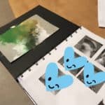 Open scrapbook with an abstract green, brown, and white painting, two photographs of a dog, and three blue hearts with smiling winky faces