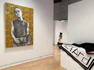Gallery view of the exhibition Presently. Fiona Crangle's mixed media on canvas featuring a person cradling a large microphone on a floral background and Brandon Wulff’s black and white quilt draped on a plinth installed in the AGP’s main gallery.