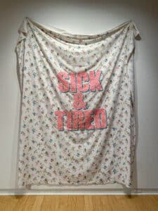 Textile by Andrew McPhail installed in the AGP's main gallery. Floral bedsheet with the words "Sick & Tired" stitched in in pink and green sequins.