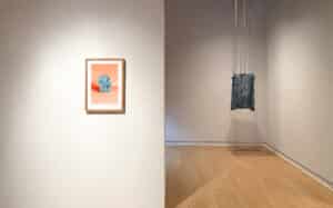 Gallery view of the exhibition Presently featuring Alison Postma's archival pigment print of a scrimped blue paper form on a peach background and Emma Schnurr's sculpture of a blue and white fabric bag suspended by industrial chain installed in APG's main gallery