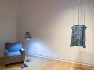 Gallery view of the exhibition Presently featuring Tonya Corkey's sculpture of a chair, lamp and slippers made with found lint on and Emma Schnurr's sculpture of a blue and white fabric bag suspended by industrial chain installed in the AGP's main gallery