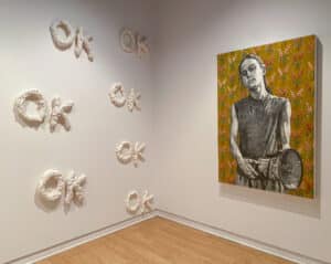 Gallery view of the exhibition Presently. Tara Lynn MacDougall's sculpture features the word "OK" repeated several times and spelt with spray insulation foam. Fiona Crangle's mixed media features a person cradling a large microphone on a floral background.