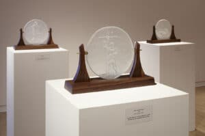 Gallery view of the exhibition La Rábida. Three glass sculptures on plinths installed in the AGP's main gallery. Etched glass featuring Cristóbal Colón, Saint Junípero Serra, La Doncella, and the maiden.