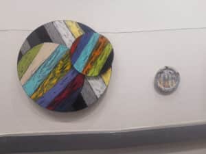 Two artworks hanging on gallery wall. Circular kinetic artwork on left and painted ceramic bowl on right