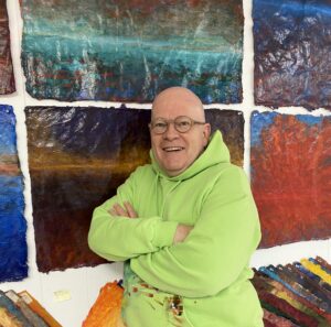 Caucasian male, bald with glasses, wearing a lime green sweater and arms crossed. He stands in front of a wall with paintings hung in a grid pattern