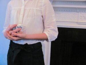 A person in a white shirt and black pants clasps a floral patterned teacup in both of their hands. The photo is cropped so that only their torso is visible